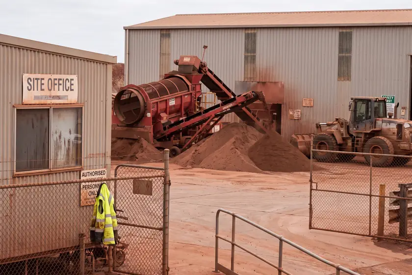 A trommel screen discharging onto a stockpile in a red dirt quarry yard, with a wheel loader and a demountable site office behind it.