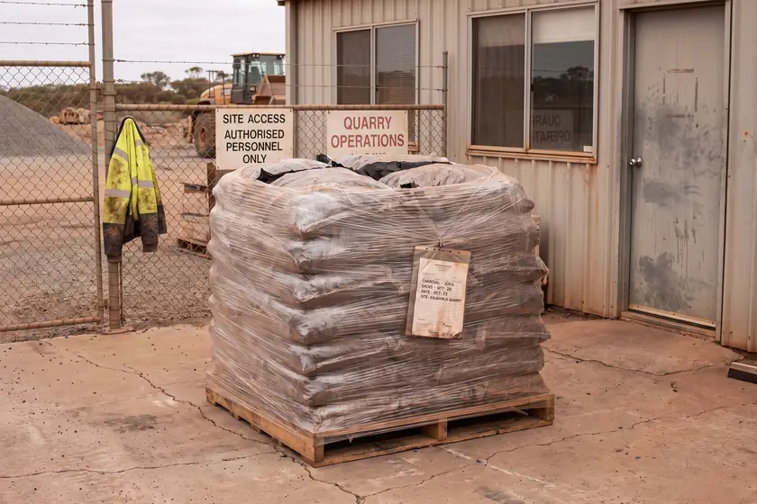 A shrink-wrapped pallet of bagged dirt standing on sealed concrete outside the site office, consignment docket taped to the wrap.
