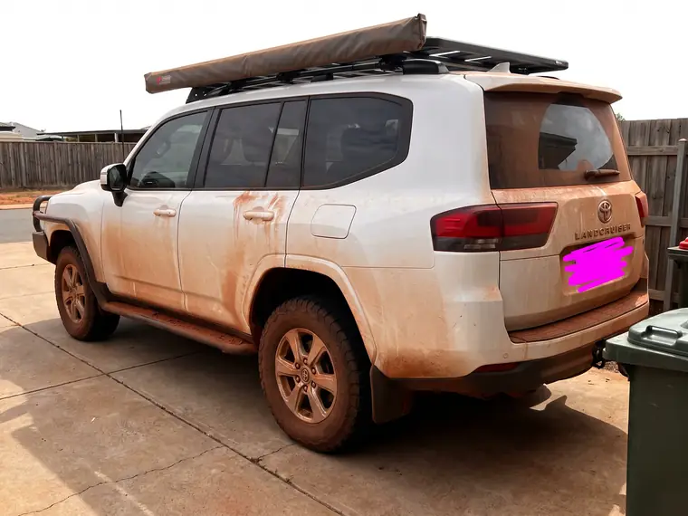 Customer photo of a white LandCruiser 300 with a roof rack and rooftop tent on a suburban driveway, red dust worked up the lower doors, rear quarter and tailgate. The number plate has been scribbled out.